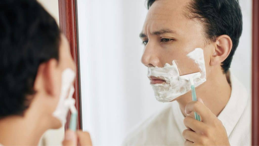 Man in a white robe shaving his face with cream in a mirror.