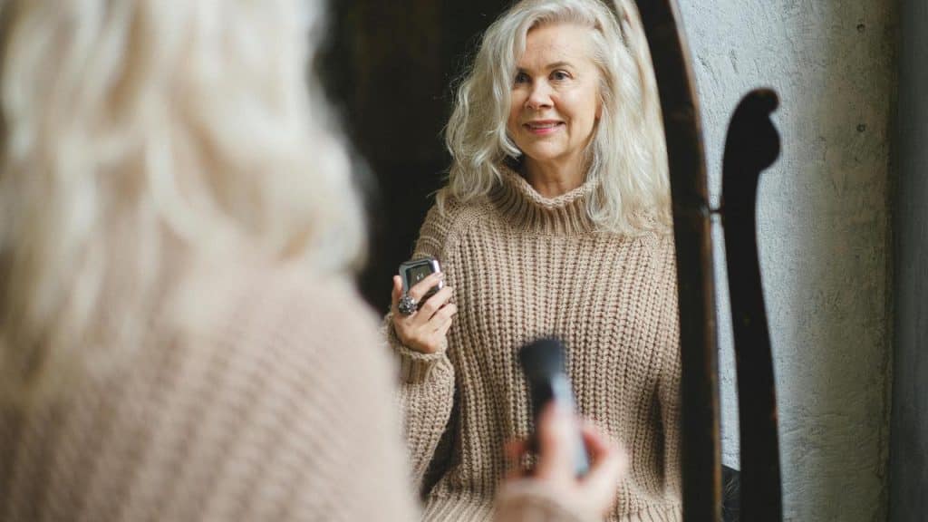 An elderly woman smiling while applying makeup and looking into a mirror.