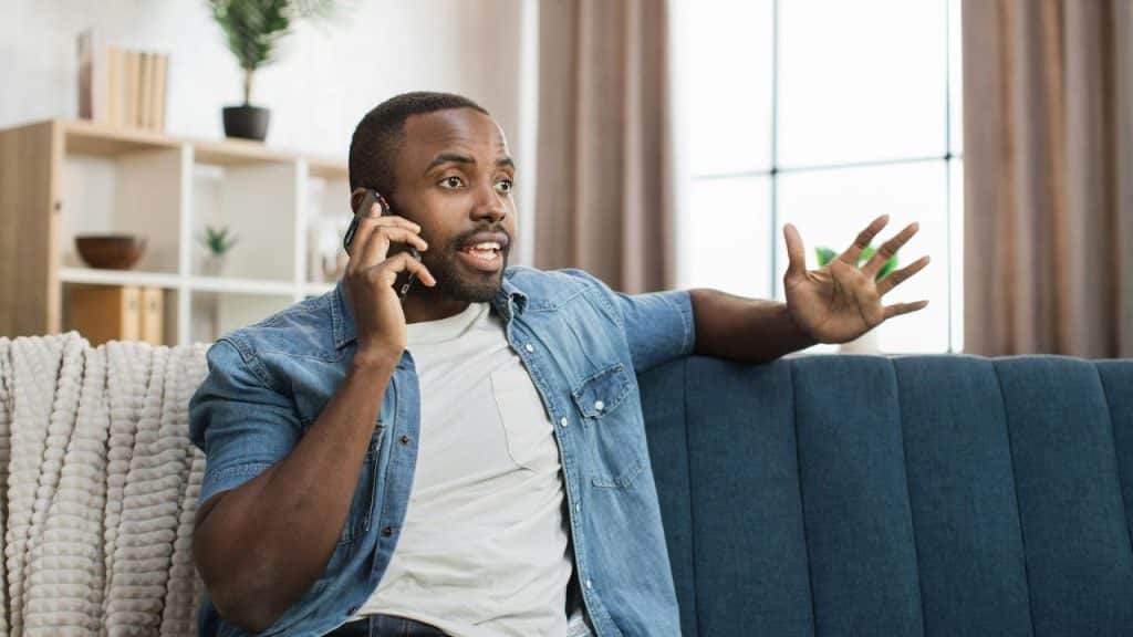Man in a denim shirt talking animatedly on a phone while sitting on a couch.