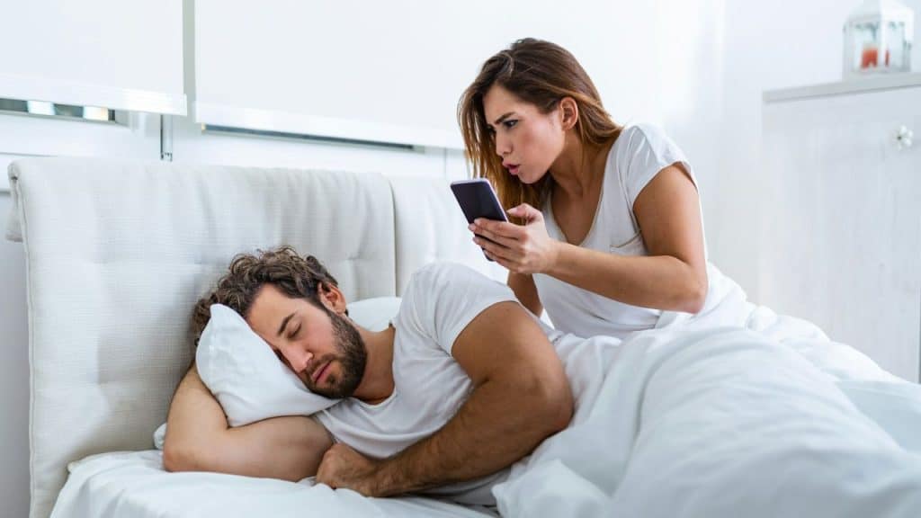 A woman secretly reads a phone while a man sleeps next to her in bed.