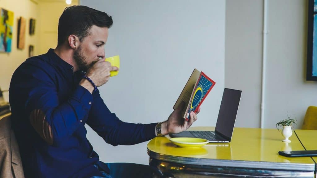 Man with a beard in a blue shirt reading a book and drinking from a yellow mug at a yellow table with a laptop.