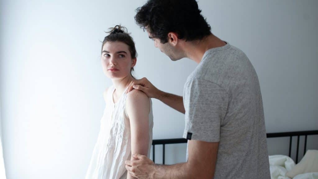 A serious-looking woman in a white nightgown is standing next to a bed while a man puts a comforting hand on her shoulder.