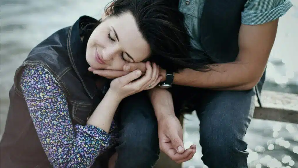 A woman resting her head peacefully on a man's hand by the water.