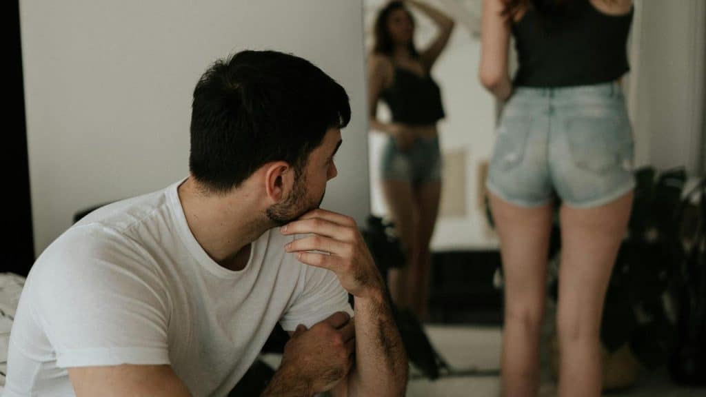 A man sitting and looking toward a woman standing in front of a mirror.