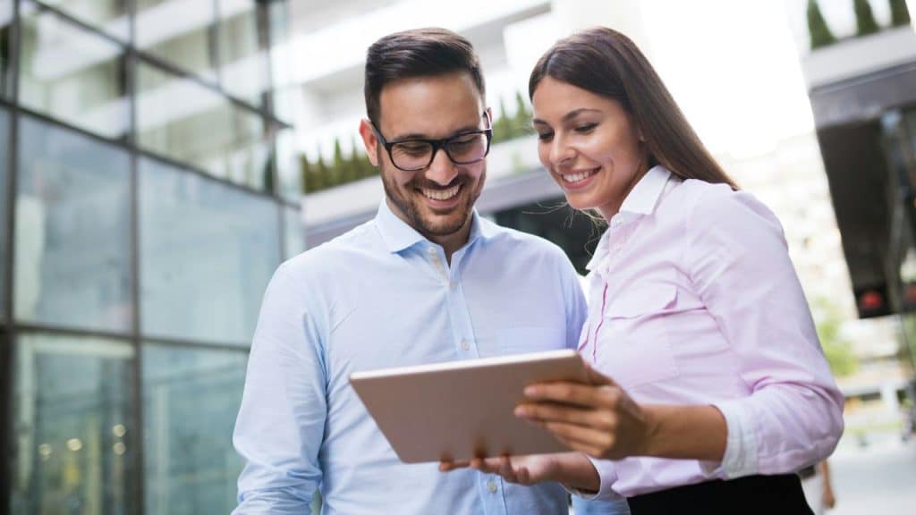 Two smiling businesspeople looking at a tablet together outdoors in front of a modern glass building.