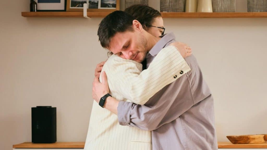 Man embracing another person who is wearing a light-colored jacket indoors.