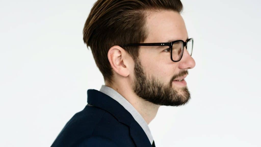 Profile close-up of a man with a beard and glasses wearing a dark jacket.