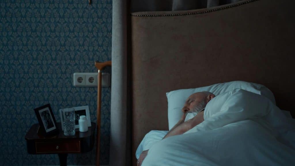 An elderly man sleeping in bed beside a nightstand with photos and a glass of water.