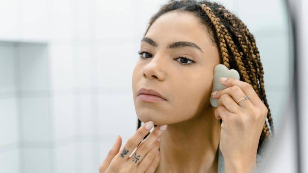 A woman uses a Gua sha tool on her face while looking in the mirror.