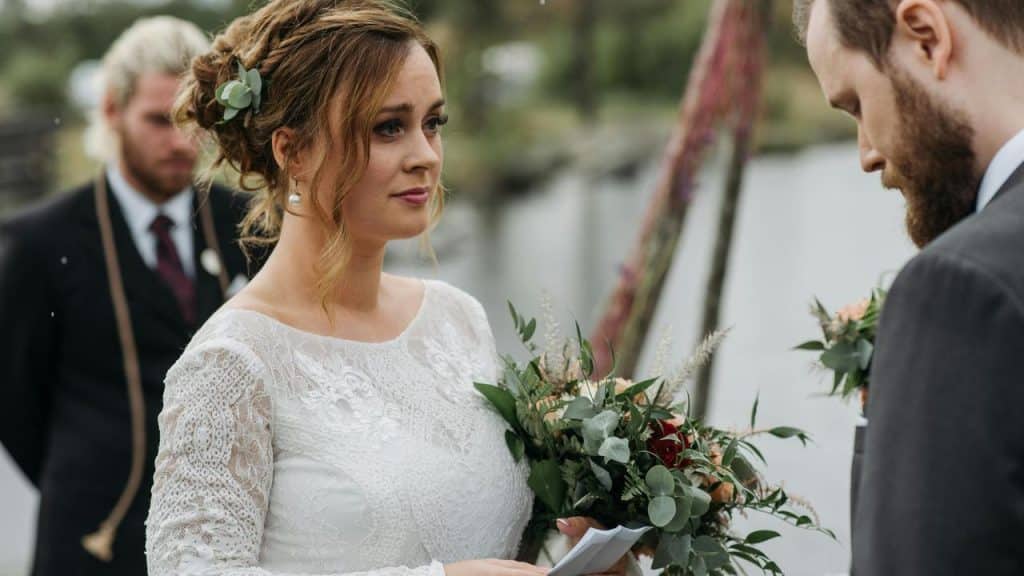 A bride holding a bouquet looks at the groom during an outdoor wedding ceremony.
