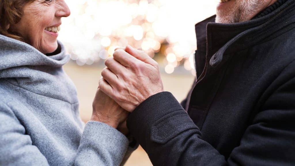 A couple in coats smiles and holds hands warmly outdoors.