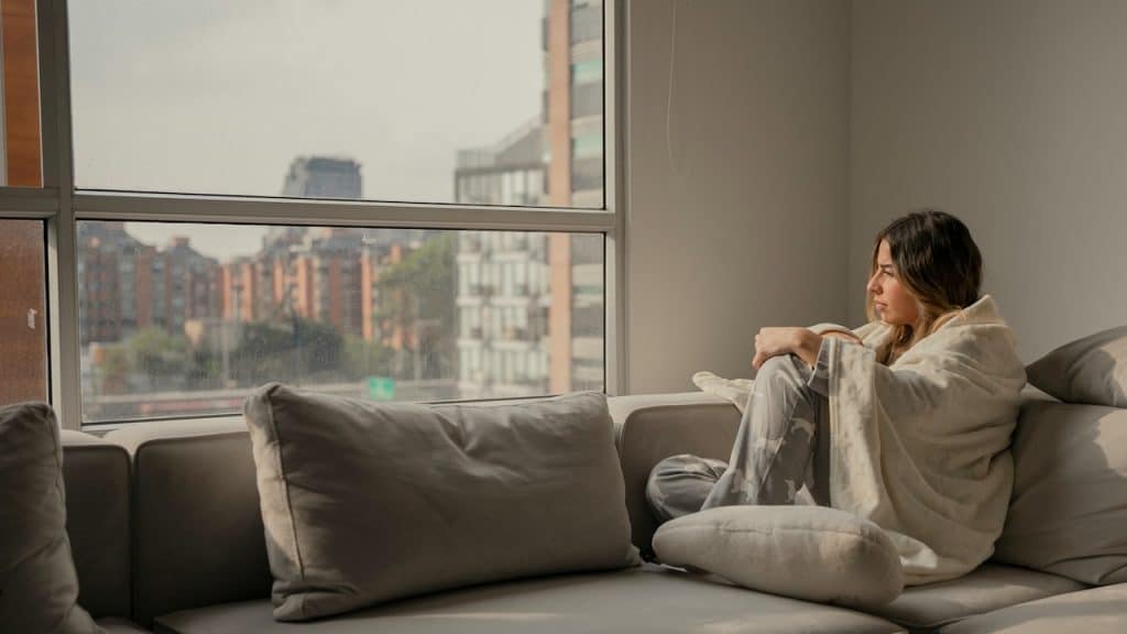 A solitary woman is sitting on a large, neutral-colored couch, wrapped in a light blanket, gazing out a window at a cityscape.