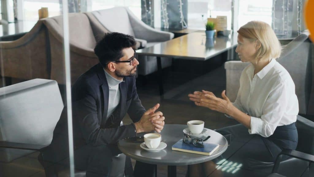 A man listening to a woman gesturing while sitting at a coffee table.