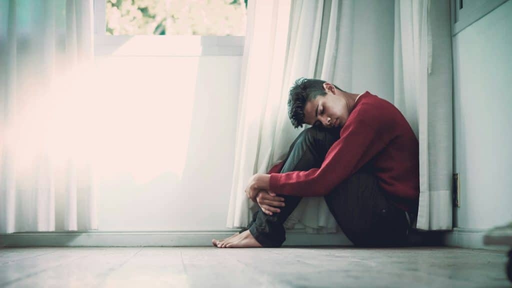 A young man in a red sweater is sitting on a wooden floor, hugging his knees to his chest, looking sad.