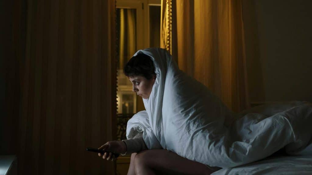 A person wrapped in a blanket sitting on a bed while watching something on a screen.