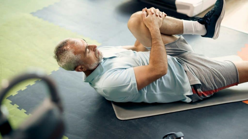 A silver-haired man lies on a mat, stretching his knee toward his chest in a gym.