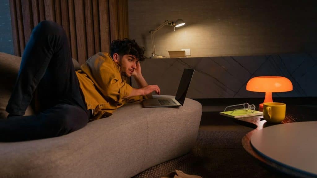 A man is lying on a sofa, looking at his laptop, with a dim light source nearby.
