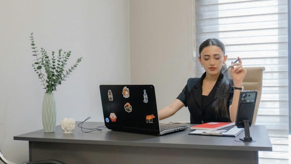 A professional young woman sitting at an office desk, looking at her laptop and holding a pen.