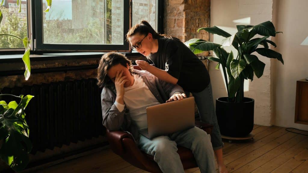 A concerned woman leans over a man sitting in a chair, working on a laptop, who is covering his face with his hand.