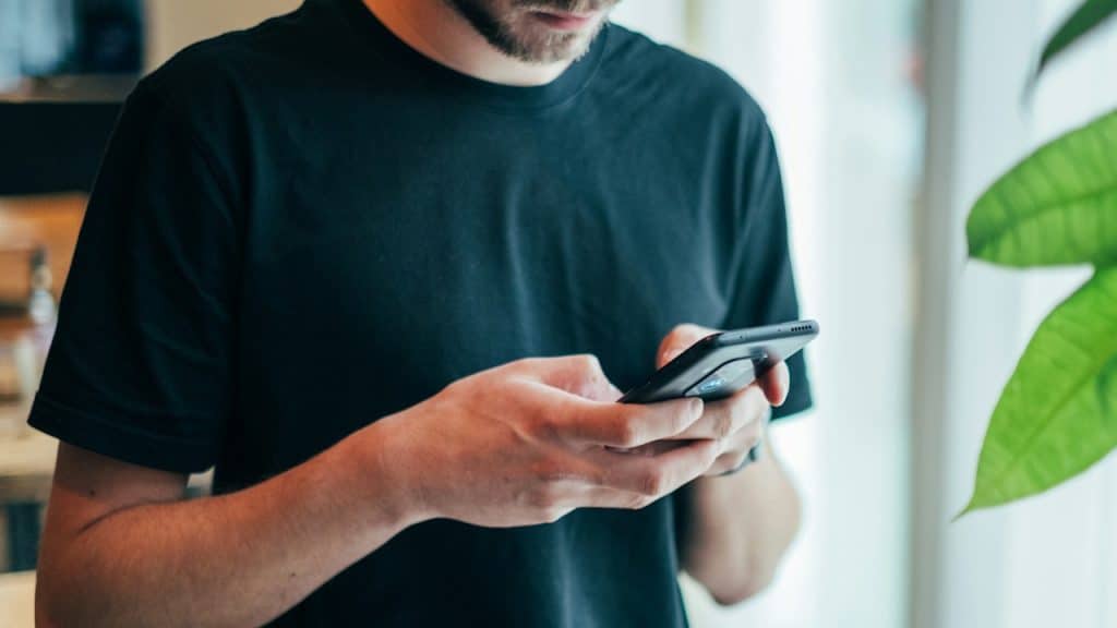 Close-up of a person in a black t-shirt looking down at and texting on a smartphone they hold with both hands.