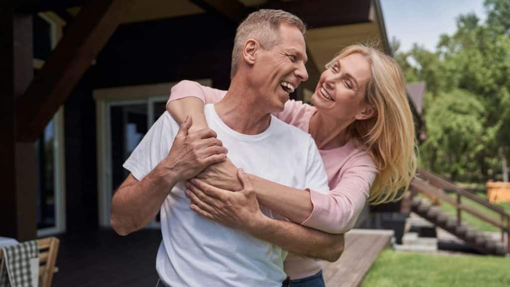 A middle-aged man and woman embrace and laugh outdoors in front of a house.