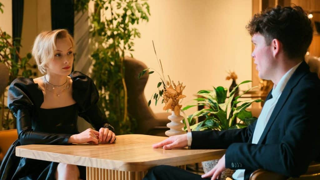 A man in a suit and a woman in a black dress sit seriously across a table.