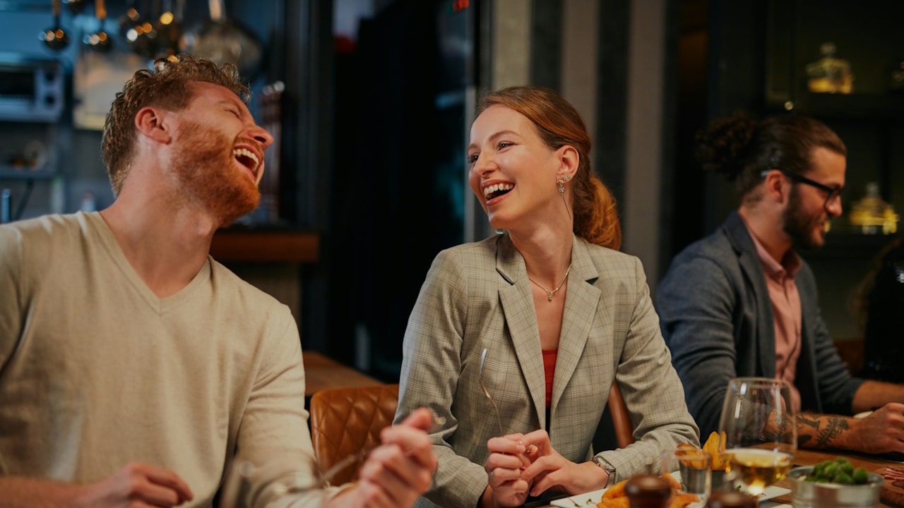 A joyful couple at dinner, with a red-haired man laughing heartily and a smiling woman in a blazer looking at him.
