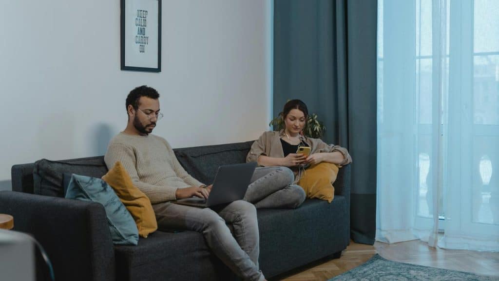 A couple sitting on a couch using a laptop and a phone.