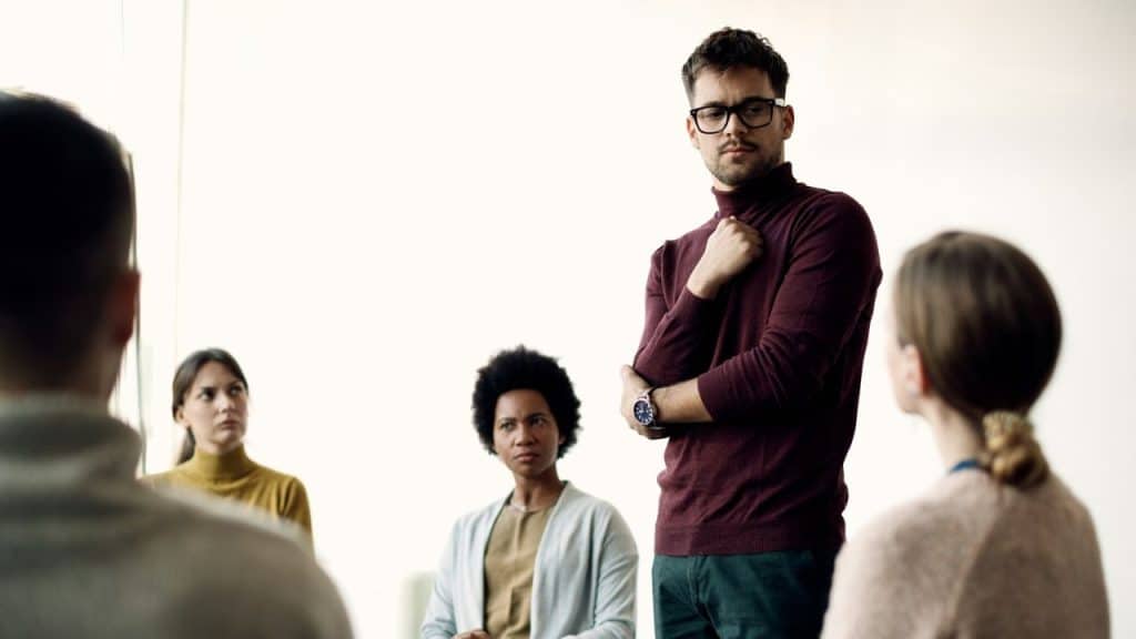 Man in a maroon turtleneck standing before a small, seated group in a bright room.