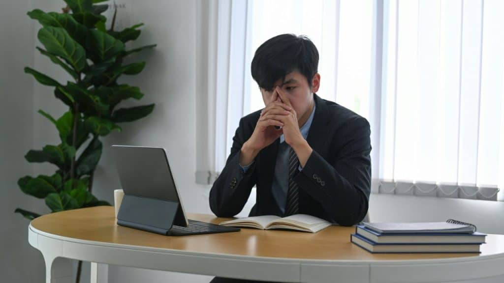 Young man in a dark suit sitting at a desk with his hands clasped over his mouth, looking stressed.