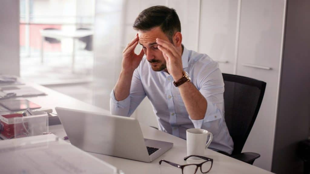 Stressed man sitting at a desk with a laptop, holding his temples in an office.