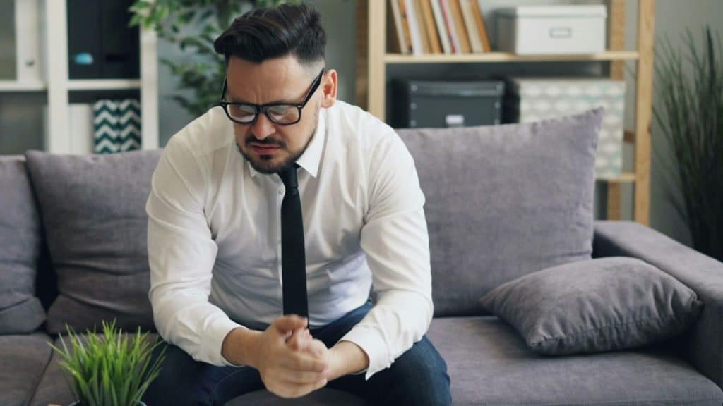 A distressed man in a white shirt and black tie sits on a couch, holding his hands together.