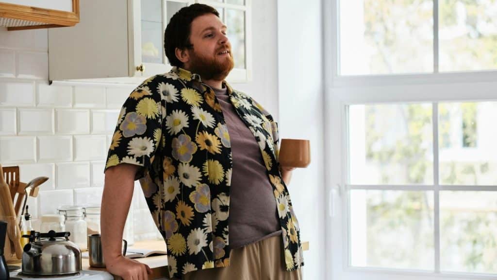 Man in a floral shirt holding a wooden mug, standing in a brightly lit kitchen.