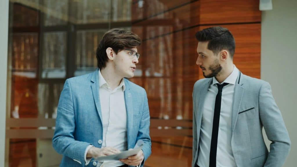 Two men in suits intensely talking face-to-face in a modern glass office building.