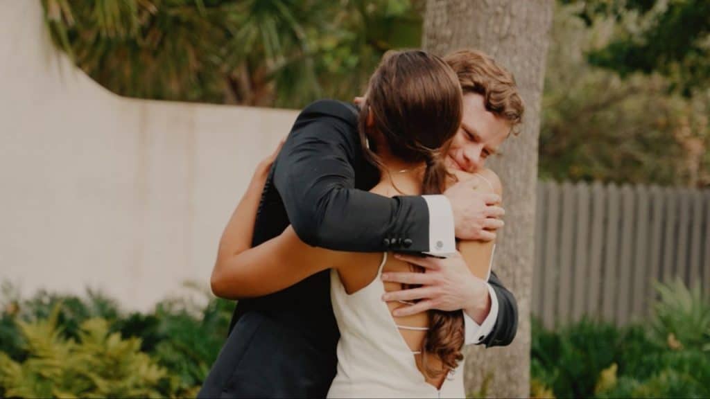Man in a black suit hugging a woman in a white dress outdoors near a tree.