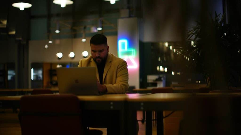 Man in a tan jacket working on a laptop in a dimly lit modern office space.