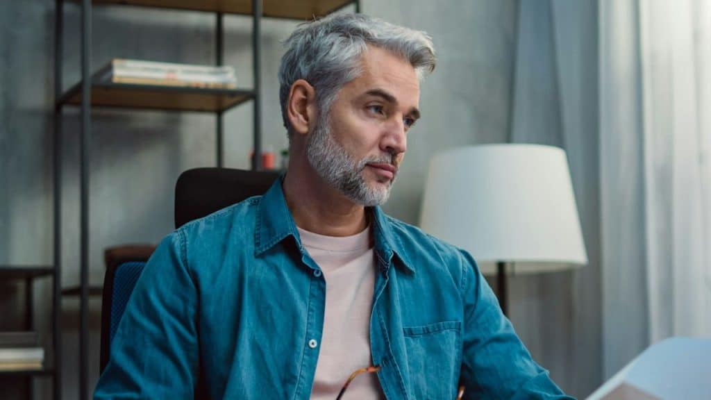 A middle-aged man with gray hair and a beard sits at a desk looking thoughtful.