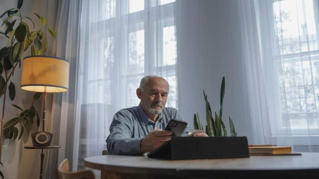 An older man using both a smartphone while seated at a softly lit table.