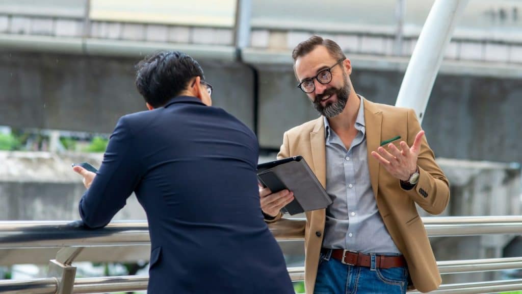 Bearded man holding a tablet and gesturing while talking to another man outdoors.