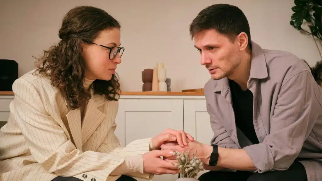 Man and woman facing each other indoors, holding hands and looking serious.