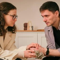 Man and woman facing each other indoors, holding hands and looking serious.