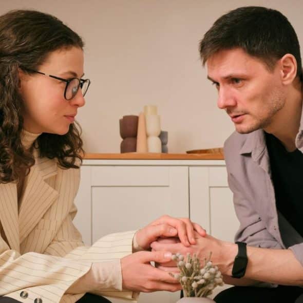 Man and woman facing each other indoors, holding hands and looking serious.
