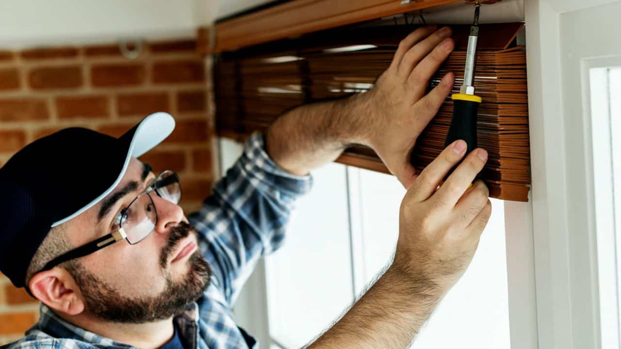 Man in a cap and glasses using a screwdriver to install wooden blinds near a window.