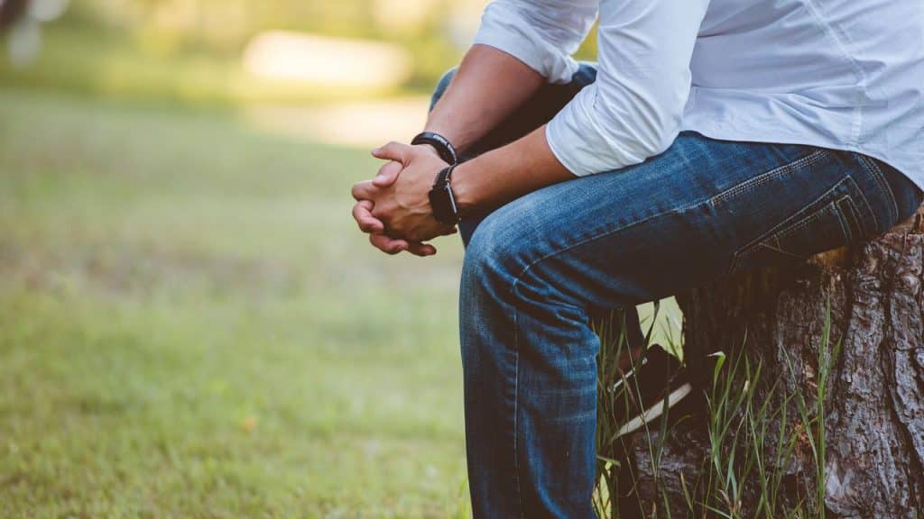 A person sitting on a tree stump outdoors with hands clasped and head out of frame.