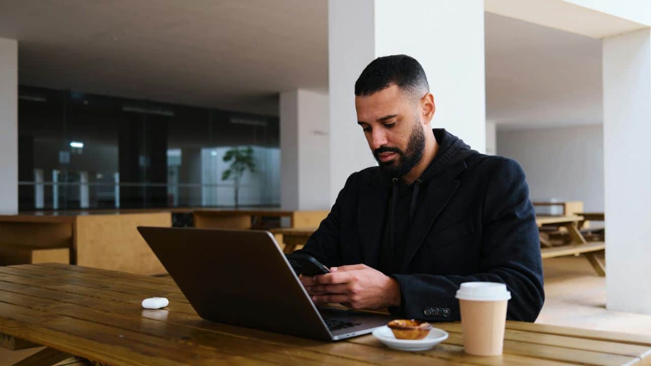 Bearded man using a smartphone at a wooden outdoor table with a laptop and coffee.
