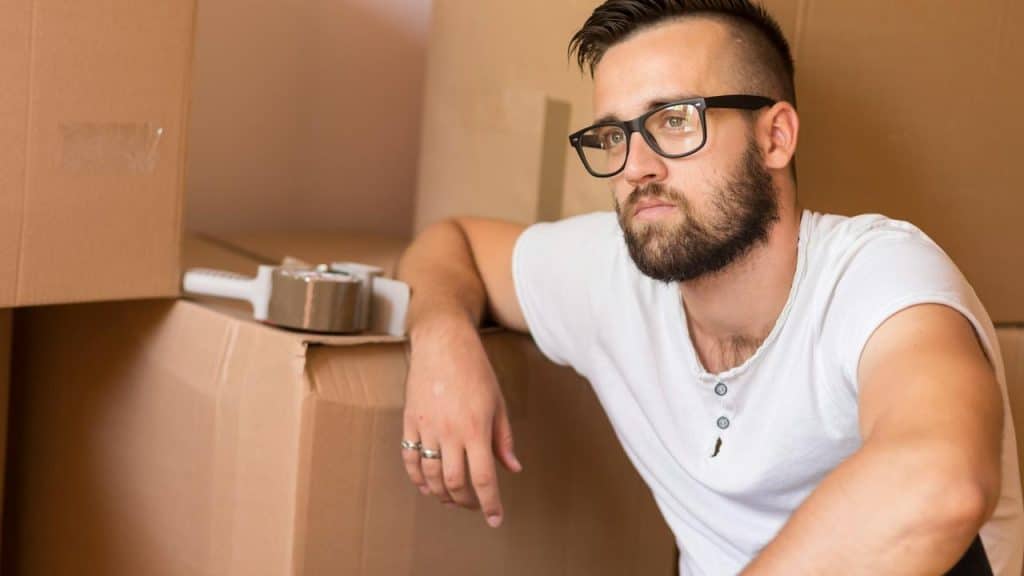A man with glasses leans against stacked moving boxes, looking thoughtful.