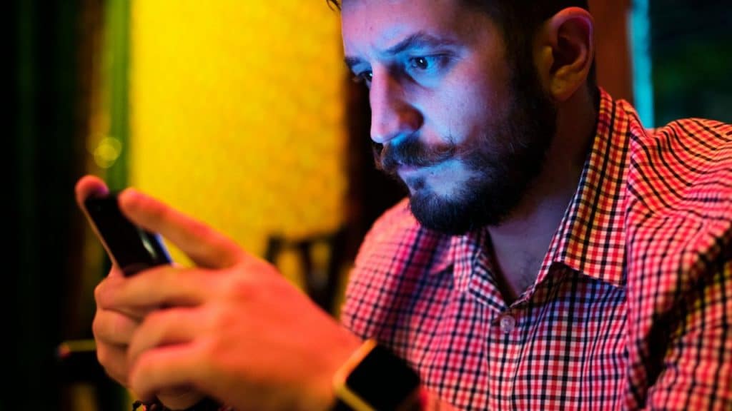 Man with a mustache and beard looking at his phone in dimly lit, colorful light.