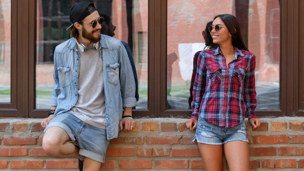 A couple wearing sunglasses standing alongside each other while leaning on a wall.