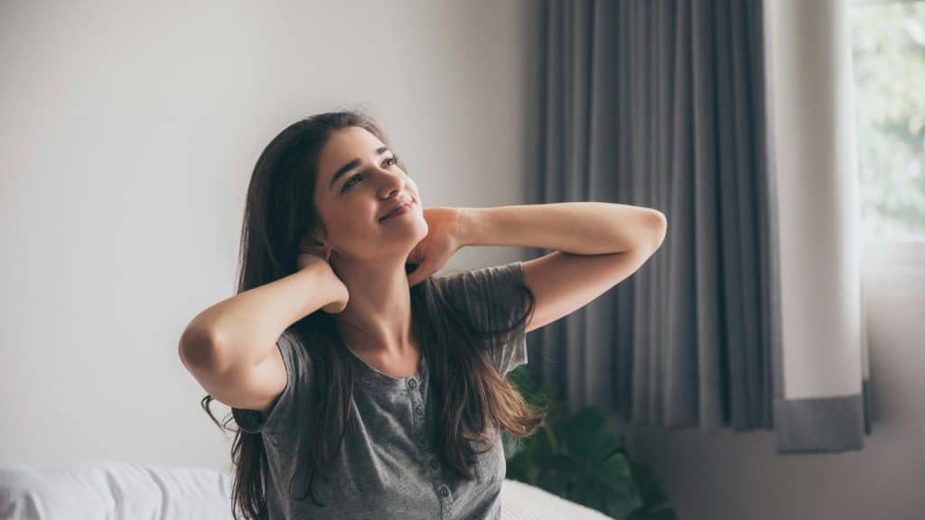 A woman sits on a bed smiling and stretching with her hands on her neck.