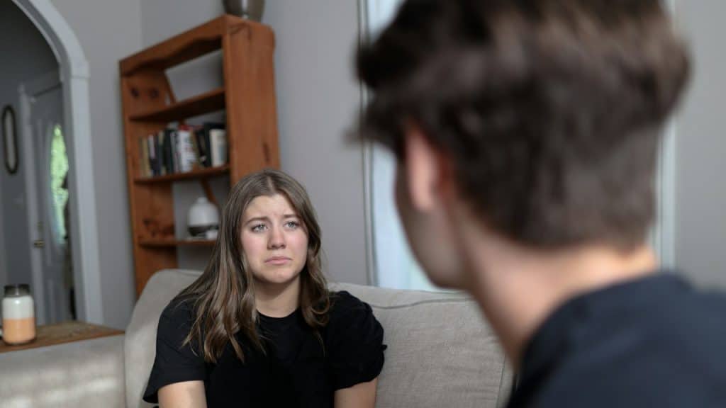 A woman looking at a man while sitting on a couch with a sad expression on her face.